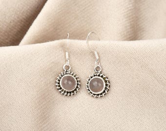 May include: A pair of silver-toned earrings with a round, light pink stone center. The earrings have a decorative, twisted rope-like border. The earrings are displayed on a beige fabric background.