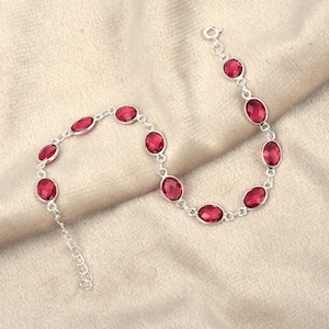 以下が含まれることがあります： 深い赤色の楕円形の宝石をあしらったシルバーブレスレット。宝石はチェーンにセットされており、繊細でエレガントなデザインです。ブレスレットはニュートラルカラーの生地の上に展示されています。