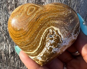 May include: A heart-shaped stone with a polished surface. The stone features swirling patterns in shades of brown, tan, and white. The heart is held in a hand with light blue nail polish. The stone appears to be a decorative item or a natural specimen.
