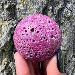 May include: A pink, round, porous, and textured gemstone sphere. The sphere has a rough, uneven surface with many small holes.