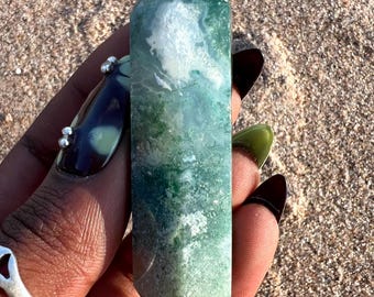 May include: A green and white agate crystal point, also known as a wand, with a double-terminated point. The crystal is held in a hand.