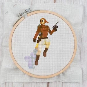 May include: Cross-stitch embroidery of a figure in a brown aviator jacket, white pants, and brown boots, holding a black gun. The figure is set against a white fabric background, framed by a wooden embroidery hoop. The design includes a light blue and purple cloud-like element.