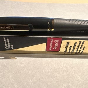 May include: A black Sanford PhD Gold mechanical pencil with gold-plated accents and an ergonomic triangular grip. The pencil is retractable and has a 0.5mm lead with a twist erase. The packaging is visible.