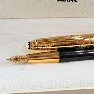 May include: A gold fountain pen with detailed engravings and a black body. The cap is off, showing the nib. The pen is in a white box. The design suggests a high-end writing instrument.