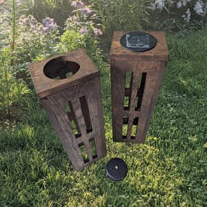 Puede incluir: Dos postes de luz solar de exterior de madera con un panel solar negro en la parte superior de cada poste. Los postes están en un jardín con hierba verde y flores.