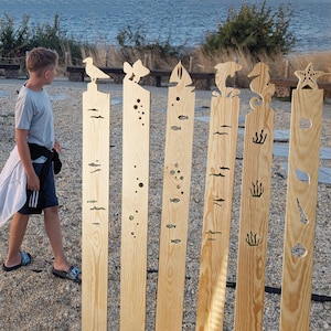 Puede incluir: Seis postes decorativos de madera con recortes de temática marina. Cada poste presenta una criatura o elemento marino diferente, incluyendo una gaviota, un pez, un velero, un delfín, un caballito de mar y una estrella de mar. Los postes son de madera clara.