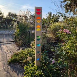 Może przedstawiać: Drewniany słupek z kolorowymi szklanymi kółkami przymocowanymi do niego. Kółka są ułożone w tęczę, z czerwonym, pomarańczowym, żółtym, zielonym, niebieskim i fioletowym. Słupek stoi w otoczeniu ogrodu z zielonymi roślinami i kwiatami.