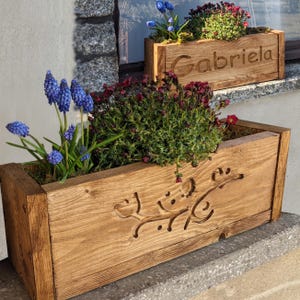 Könnte beinhalten: Zwei rechteckige Holzpflanzkästen gefüllt mit bunten Blumen. Der größere Pflanzkasten hat ein geschnitztes Design und blaue Blumen. Der kleinere Pflanzkasten hat den Namen "Gabriela" in das Holz geschnitzt. Beide stehen auf einer Betonoberfläche.