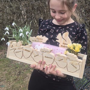 Könnte beinhalten: Eine Holzkiste mit eichelförmigen Ausschnitten, die jeweils einen Namen tragen. Die Kiste enthält Pflanzen, ein verpacktes Geschenk und Holzausschnitte mit Text. Das Mädchen hält die Kiste.
