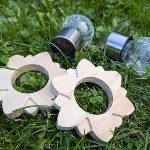 May include: Two wooden rings with a flower-like design, each with a hole in the center. The rings are on a bed of green grass. Two glass globe solar lights are also visible in the image.