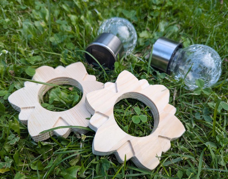 May include: Two wooden rings with a flower-like design, each with a hole in the center. The rings are on a bed of green grass. Two glass globe solar lights are also visible in the image.