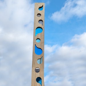 Könnte beinhalten: Ein vertikales Holzobjekt mit heller Holzmaserung. Es hat eine Reihe von ausgeschnittenen Formen, darunter Tropfen und Kreise, die mit Blau und anderen Farben gefüllt sind. Das Kunstwerk befindet sich vor dem Hintergrund eines bewölkten Himmels.