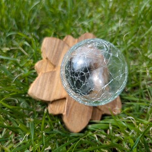 May include: A clear glass sphere with a crackled surface sits on a wooden base shaped like a flower. The sphere is resting on a bed of green grass.
