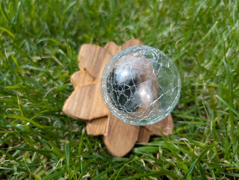 May include: A clear glass sphere with a crackled surface sits on a wooden base shaped like a flower. The sphere is resting on a bed of green grass.