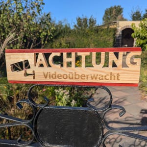 Può includere: Un cartello di legno con strisce rosse e bianche in alto e le parole "ACHTUNG Videoüberwacht" in tedesco. Il cartello è montato su un cancello di metallo nero.