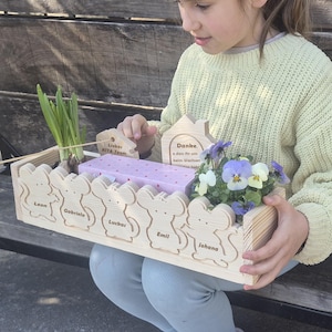 Könnte beinhalten: Ein hölzerner Geschenkkorb mit dekorativem Mausdesign, der eine kleine Pflanze, ein verpacktes Geschenk und Holzzuschnitte mit Text enthält. Die Namen "Leon", "Gabriela", "Luchas", "Emil" und "Johana" sind auf dem Korb eingraviert.