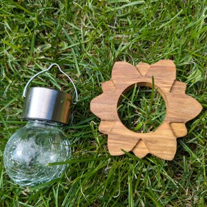 May include: A small, clear glass globe solar light with a silver metal top and a wire handle. The light is sitting on a bed of green grass next to a wooden, flower-shaped ring.