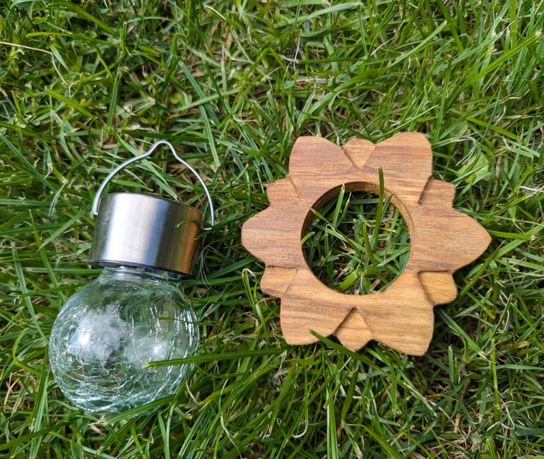 May include: A small, clear glass globe solar light with a silver metal top and a wire handle. The light is sitting on a bed of green grass next to a wooden, flower-shaped ring.