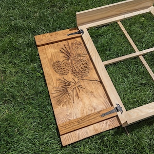 Könnte beinhalten: Ein Holzklappen mit einem geschnitzten Kiefernzapfen-Design. Die Klappe ist geschlossen und hat zwei schwarze Metallscharniere.