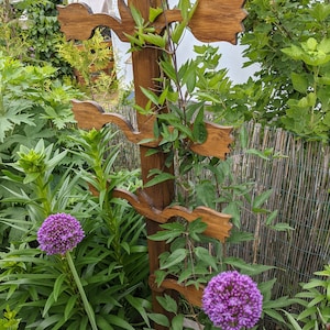 Pode incluir: Um treliça de madeira com um design decorativo que se assemelha a flores estilizadas. A treliça é coberta por videiras verdes e tem duas cabeças de flores roxas em primeiro plano.
