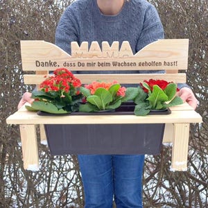 Könnte beinhalten: Ein hölzerner Bank-Stil-Pflanzkasten mit dem Wort "MAMA" und deutschem Text. Der Pflanzkasten enthält drei Topfpflanzen mit roten und orangefarbenen Blüten. Die Bank wird von einer Person gehalten, die einen grauen Pullover und Jeans trägt.