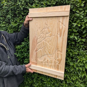 Op de afbeelding: Houten wandkunst met een uitgesneden bosscène, inclusief een vos, bomen en vogels. Het kunstwerk is gemaakt van lichtgekleurd hout met een natuurlijk nerfpatroon. De vos wordt afgebeeld in een rennende pose.