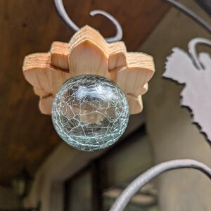 May include: A light blue glass orb with a crackled texture hangs from a wooden flower-shaped ornament. The ornament is attached to a black metal frame.