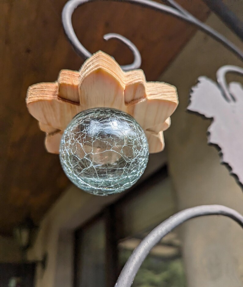 May include: A light blue glass orb with a crackled texture hangs from a wooden flower-shaped ornament. The ornament is attached to a black metal frame.