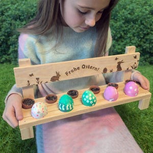 Könnte beinhalten: Eine Holzbank mit dem Text "Frohe Ostern!" und österlichen Ausschnitten. Die Bank hält kleine Töpfe und bunte Ostereier. Die Eier sind mit verschiedenen Mustern und Farben verziert, darunter Grün, Rosa und Weiß.