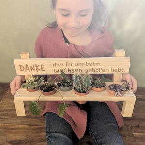 Könnte beinhalten: Eine Holzbank mit den Worten "DANKE dass Ihr uns beim Wachsen geholfen habt."  eingegraben auf der Oberseite. Die Bank hält sechs kleine Topfpflanzen, darunter Sukkulenten und Kakteen.