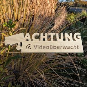 Può includere: Un cartello di legno con la parola "ACHTUNG" e il testo "Videoüberwacht" sotto. Il cartello è a forma di telecamera di sicurezza.