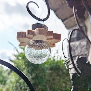 May include: A wooden light fixture with a star-shaped base and a clear glass bulb with a textured surface. The fixture is hanging from a black metal hook.