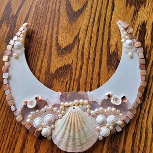 May include: A white crescent moon-shaped piece of wood decorated with seashells, pearls, and other ocean-themed embellishments. The center of the piece features a large scallop shell.