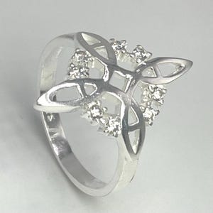 May include: A silver ring with a Celtic knot design. The ring features a central knot motif adorned with small, clear gemstones. The band is smooth and polished, and the ring is set against a light background.