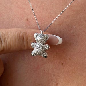 Pode incluir: Um colar de prata com um pingente de urso de pelúcia. O pingente é cravejado com pequenas pedras brilhantes e tem um detalhe em forma de coração no peito. A corrente do colar é delicada e prateada.