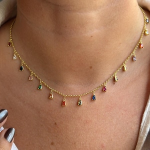Peut inclure: Un collier doré avec une chaîne fine et de petits charms en pierres précieuses multicolores. Les breloques ont différentes formes, dont des gouttelettes et des rectangles, et présentent un arc-en-ciel de couleurs. Le collier est porté autour du cou.