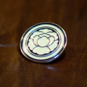Puede incluir: Un pin de esmalte blanco y azul con un diseño de flor estilizado. El pin es circular y tiene un acabado de metal plateado.