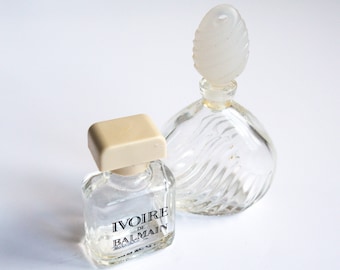 May include: Two clear glass perfume bottles with white plastic stoppers. The smaller bottle has the text "Ivoire de Balmain" on the label.