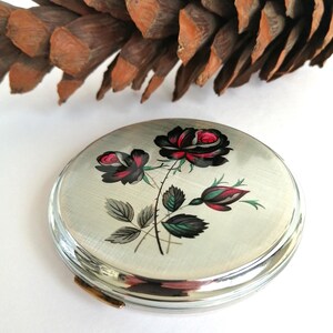 May include: A silver compact with a floral design featuring black and red roses. The compact has a clear, rounded edge and a gold clasp. A pine cone is in the background.