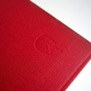 May include: Close-up of the red cover of a hardcover book. The cover has a subtle textured pattern and a small, embossed design in the center.