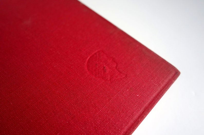 May include: Close-up of the red cover of a hardcover book. The cover has a subtle textured pattern and a small, embossed design in the center.