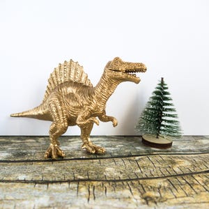 May include: A gold-painted Spinosaurus dinosaur model stands beside a small green Christmas tree on a wooden surface. The dinosaur has a large fin on its back. The tree is on a small wooden base. The scene is set on a wooden surface.