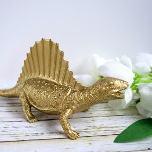 May include: A gold-painted plastic dinosaur figurine with a spiky back fin, posed on a whitewashed wooden surface with white flowers.