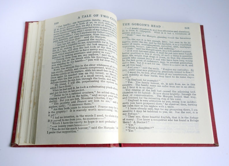 May include: An open book with a red cover. The pages are open to a chapter titled "The Gorgon's Head" from the novel "A Tale of Two Cities". The text on the page discusses the family's history and the curse of the land.
