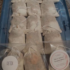 May include: A close-up of a clear plastic container filled with small, white, fabric pouches. The pouches are labeled with the text "Detox" and "Three Sisters Botanicals".