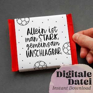 Op de afbeelding: Een witte chocolade reep verpakking met een rode rand. De verpakking heeft zwarte tekst die "Allein ist man STARK, gemeinsam UNSCHLAGBAR." luidt en een zwart-witte voetbal afbeelding.