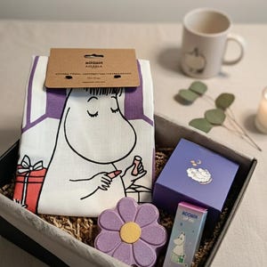 May include: A gift box featuring Moomin merchandise. It contains a white kitchen towel with a Moomin character applying lipstick, a purple flower-shaped soap, a mug, lip oil, and a purple box with a cloud design.