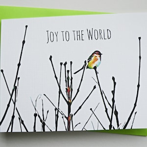 以下が含まれることがあります： 黒い文字で「JOY TO THE WORLD」と書かれた白いグリーティングカード。カードには、葉のない黒い枝のシルエットの上に止まっている小さなカラフルな鳥が描かれています。カードは明るいライムグリーンの封筒の上に置かれています。