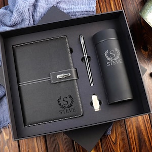 May include: A black gift set comprising a notebook, pen, tumbler, and a small accessory. The notebook and tumbler are personalised with the letter "S" and the name "STEVE". The set is presented in a black box, suitable for gifting.