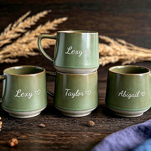 Op de afbeelding: Vier saliegroene keramische mokken met een gespikkelde afwerking en een geribbelde voet. Elke mok is voorzien van een witte naam in script en een klein hartje. De namen Lexy, Taylor en Abigail staan in een wit cursief lettertype op de mokken.